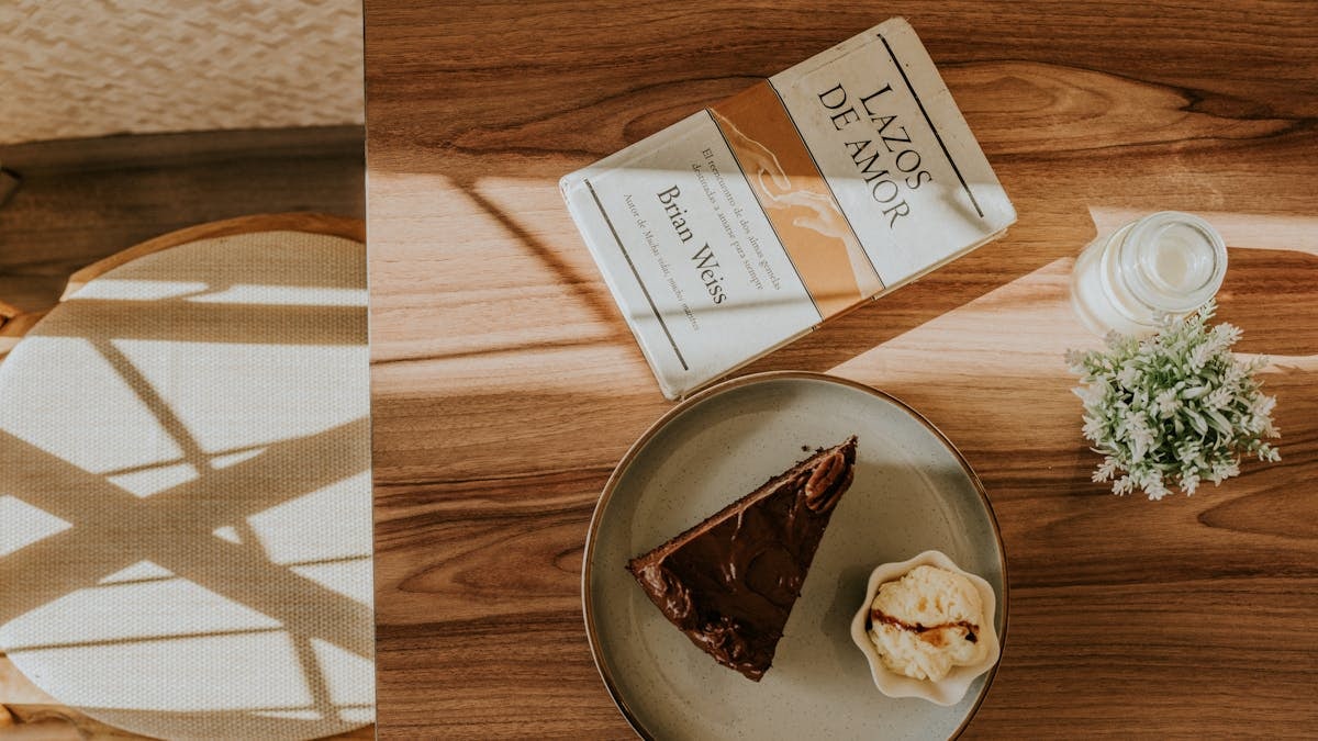 A cozy café setup with cake, ice cream, and a book on a sunlit wooden table.